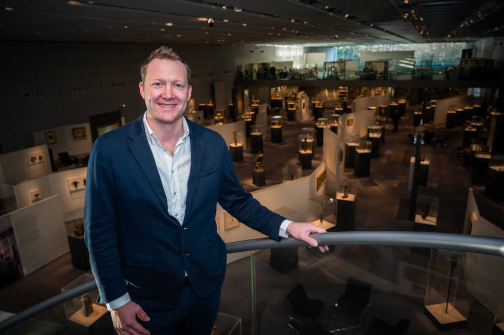 Jago Cooper A man in a suit smiling at the camera with a museum gallery in the background